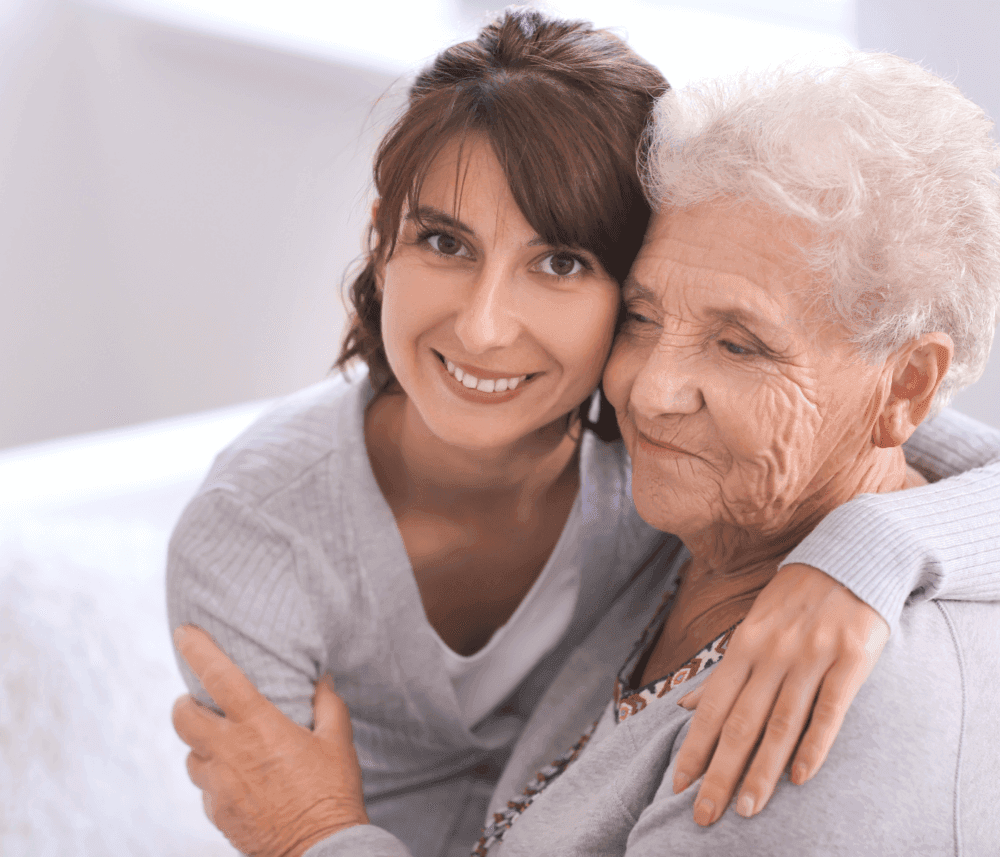 A younger woman hugs an elderly woman, both smiling warmly at the camera. - Home Instead