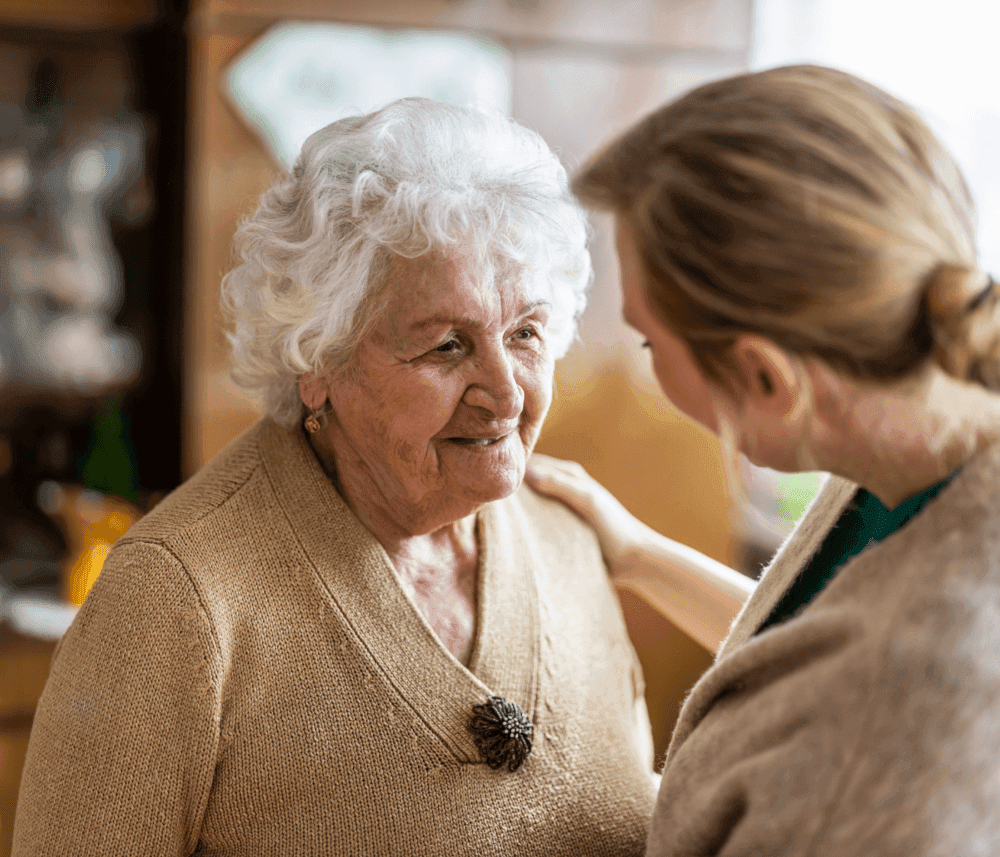 Elderly woman with white hair smiles at a younger woman who has her hand on the elderly woman's shoulder. - Home Instead