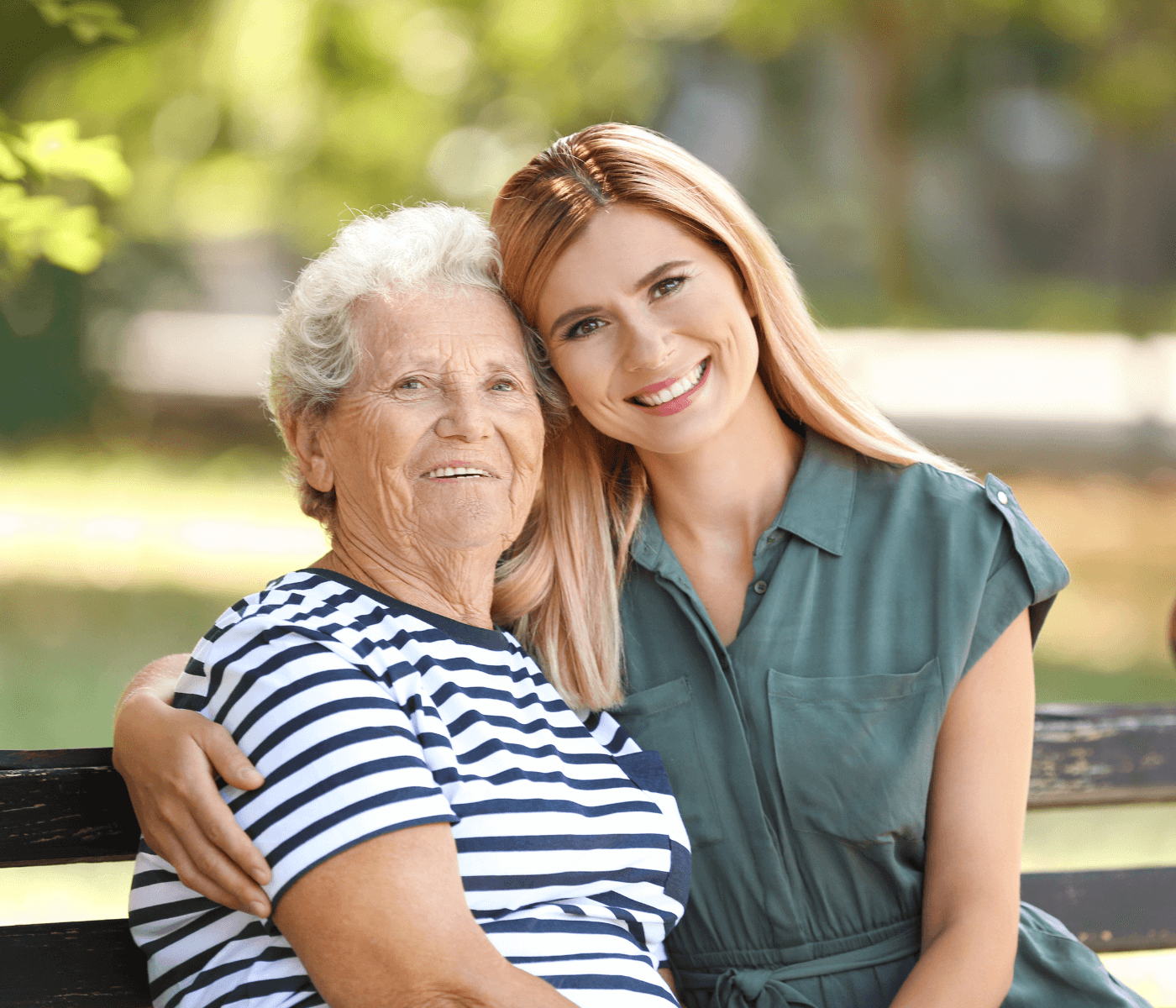 Elderly woman in a striped shirt and young woman with long hair in a green dress smile, sitting on a bench outdoors. - Home Instead