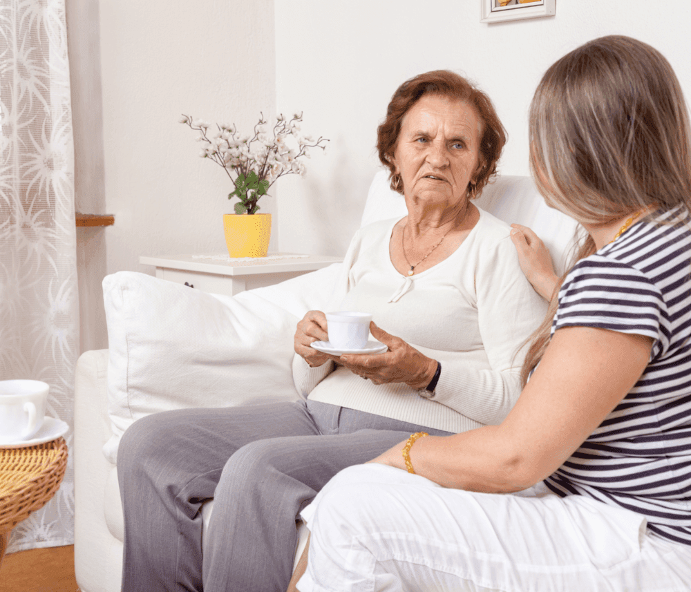 Elderly woman holding a cup, talking to a younger woman in a striped shirt, sitting together on a white couch. - Home Instead