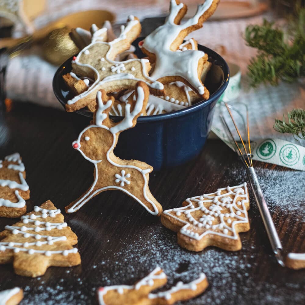 A bowl of decorated reindeer cookies with other assorted Christmas cookies on a table, surrounded by festive decor. - Home Instead