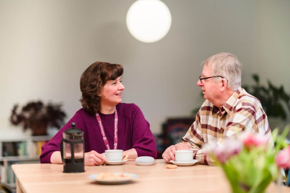 Two people sitting at a table with coffee, smiling at each other, with a blurred background and a round light above them. - Home Instead