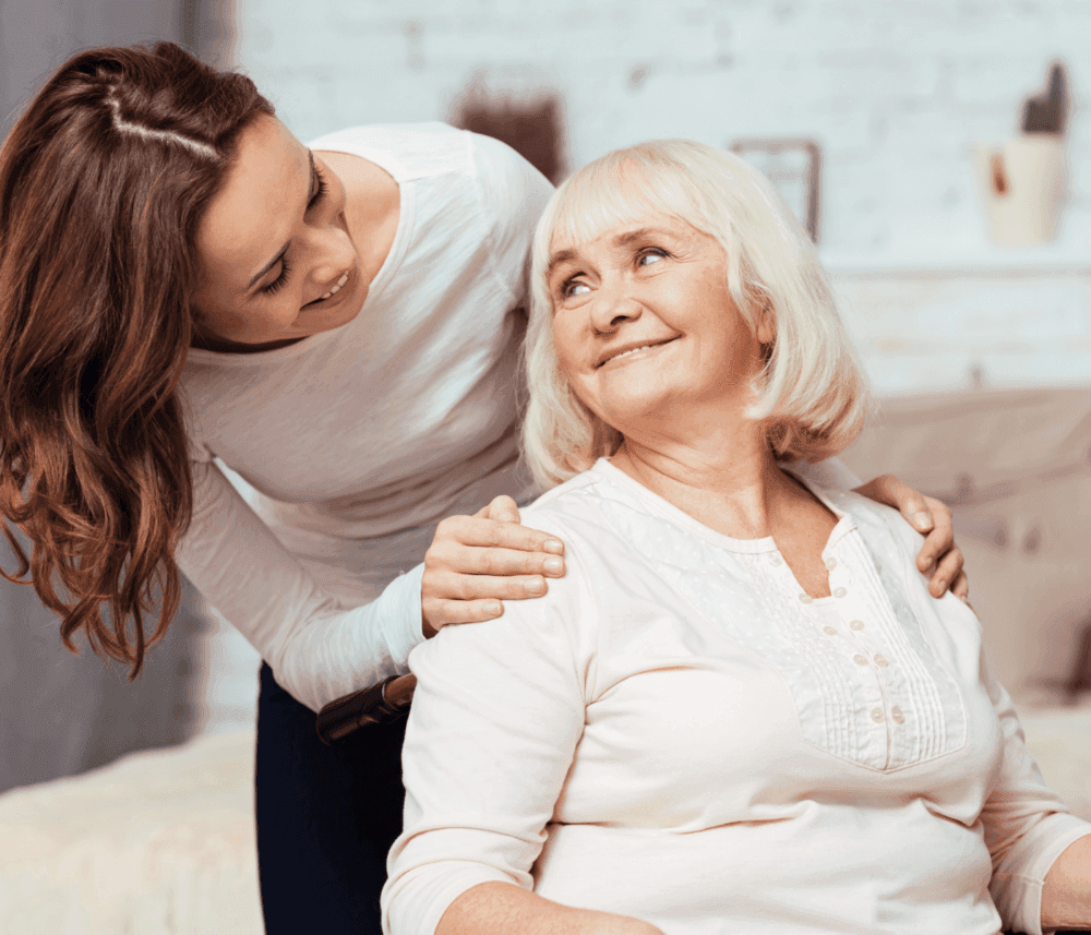 Young woman smiling at and comforting an older woman seated on a bed. Both are wearing white tops. - Home Instead