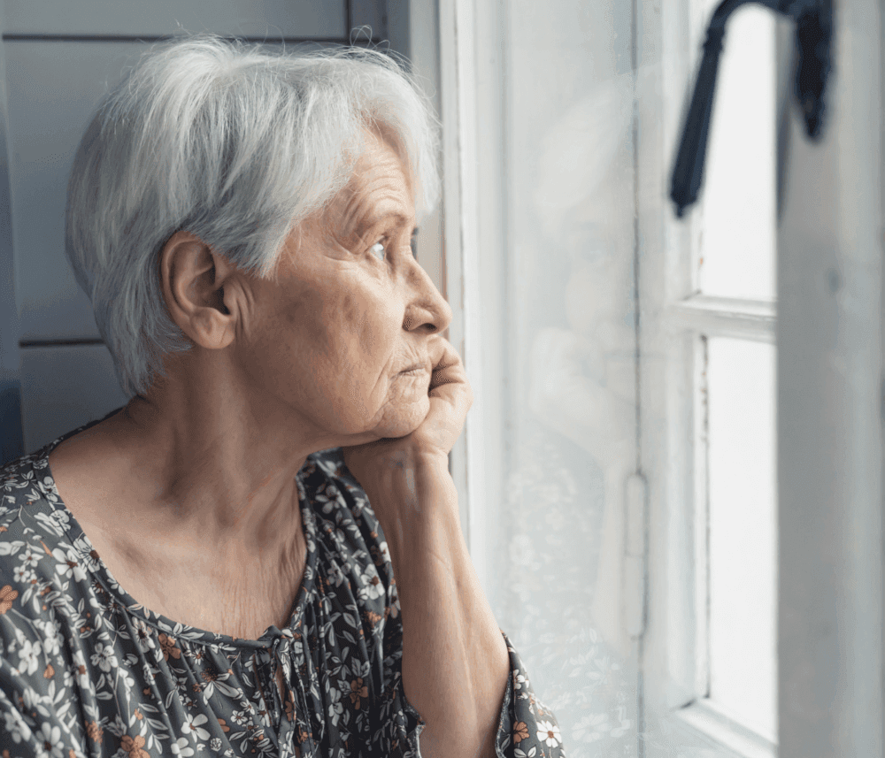 Elderly woman with short white hair, in floral blouse, looking out a window pensively, resting her chin on her hand. - Home Instead