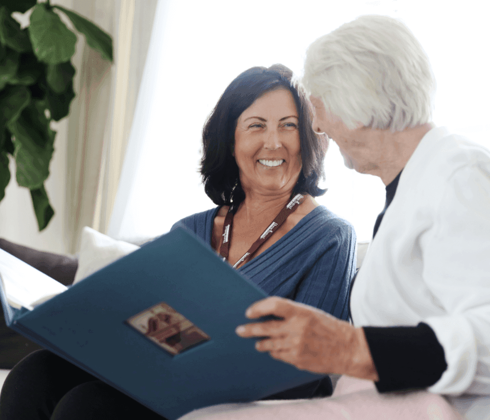 Two women smiling and looking at a large photo album together while sitting on a couch in a bright room. - Home Instead