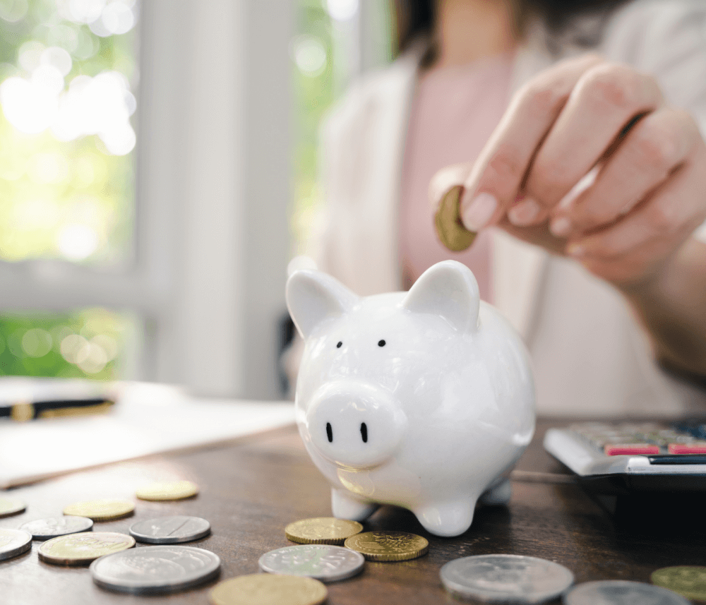 A person placing a coin into a white piggy bank, surrounded by coins and a calculator on a table. - Home Instead