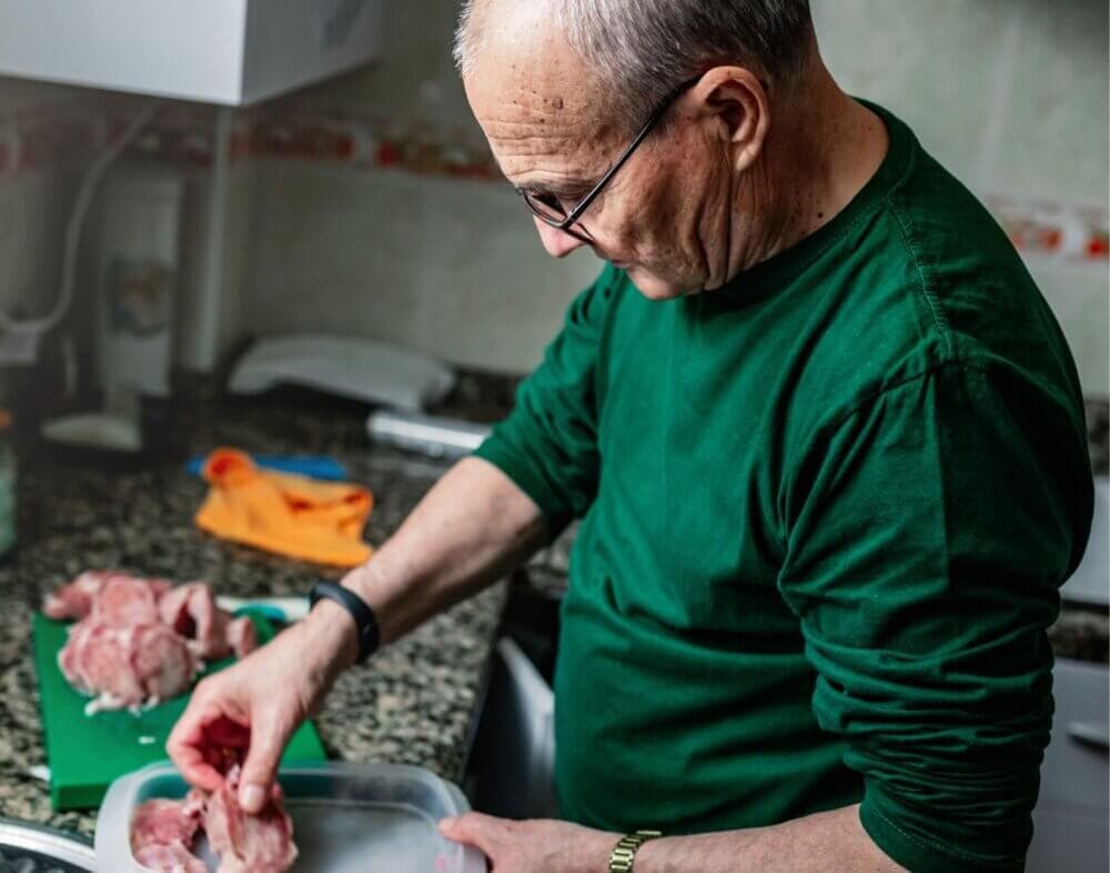 Man in a green shirt preparing raw meat in a kitchen with a granite countertop. - Home Instead