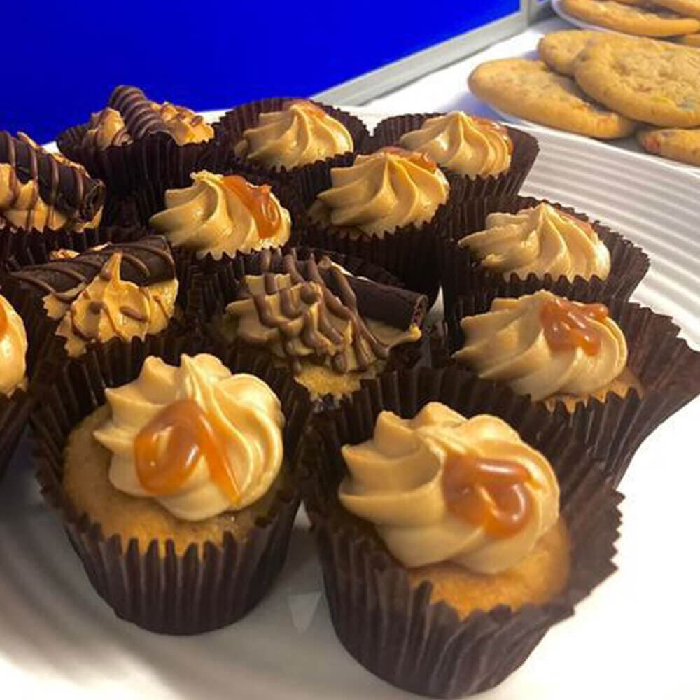 A plate of cupcakes topped with caramel and chocolate swirls, with a few cookies in the background. - Home Instead