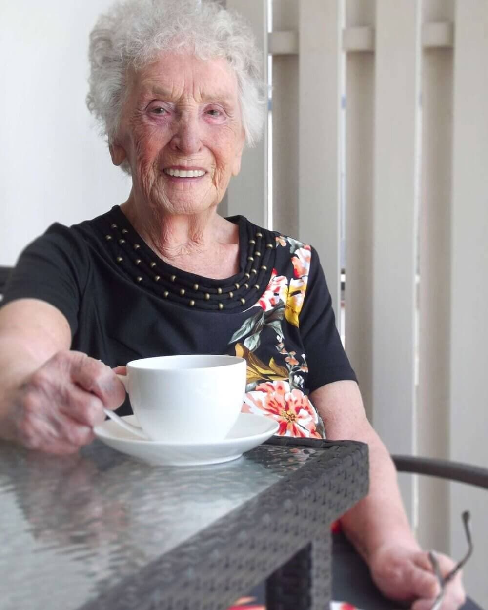 Elderly woman with white curly hair smiling, holding a white teacup, sitting at a glass table on a patio. - Home Instead