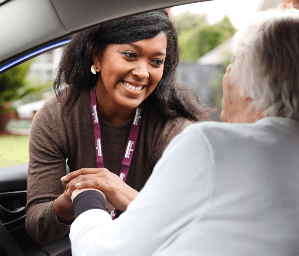 A caregiver, smiling and wearing a nametag, leans into a car to greet an older person, holding their hand. - Home Instead