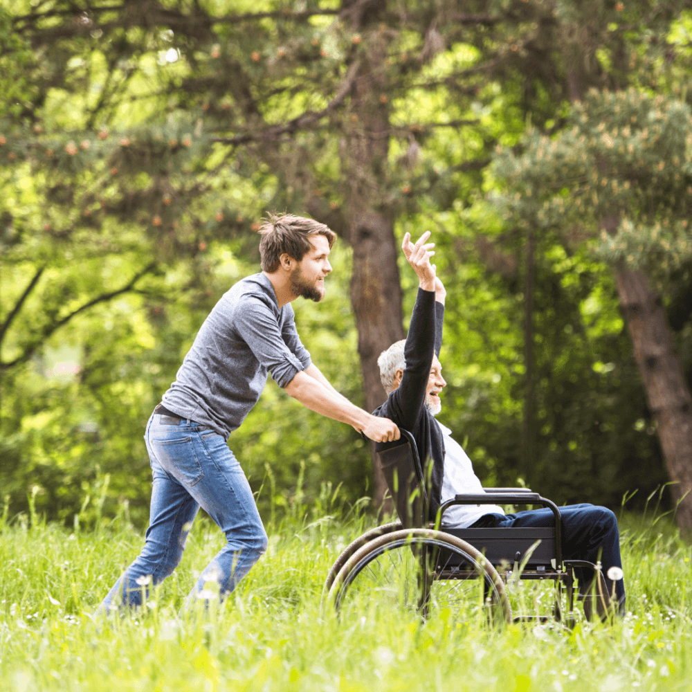 A young man pushes an elderly man in a wheelchair through a grassy, wooded area. The elderly man raises his arm. - Home Instead