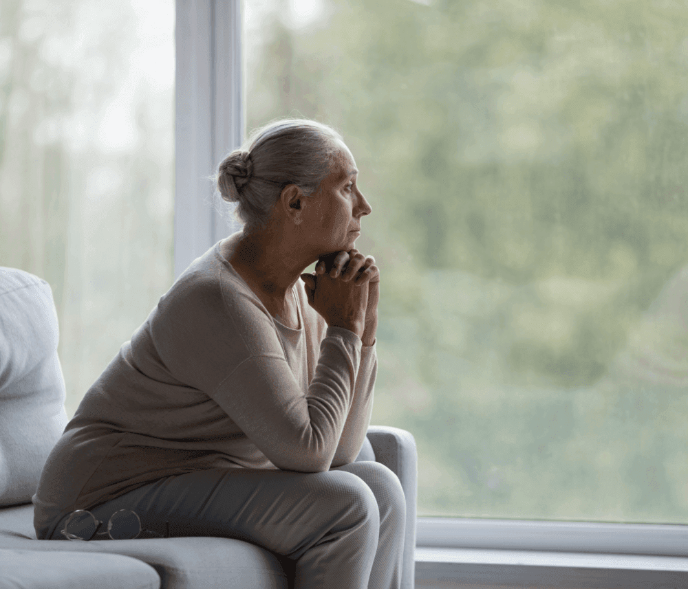 Elderly woman with gray hair sitting on a sofa, looking thoughtfully out the window with her chin resting on her hands. - Home Instead