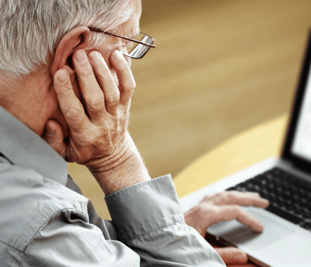 An elderly person with glasses, resting their hand on their face, uses a laptop while sitting at a table. - Home Instead