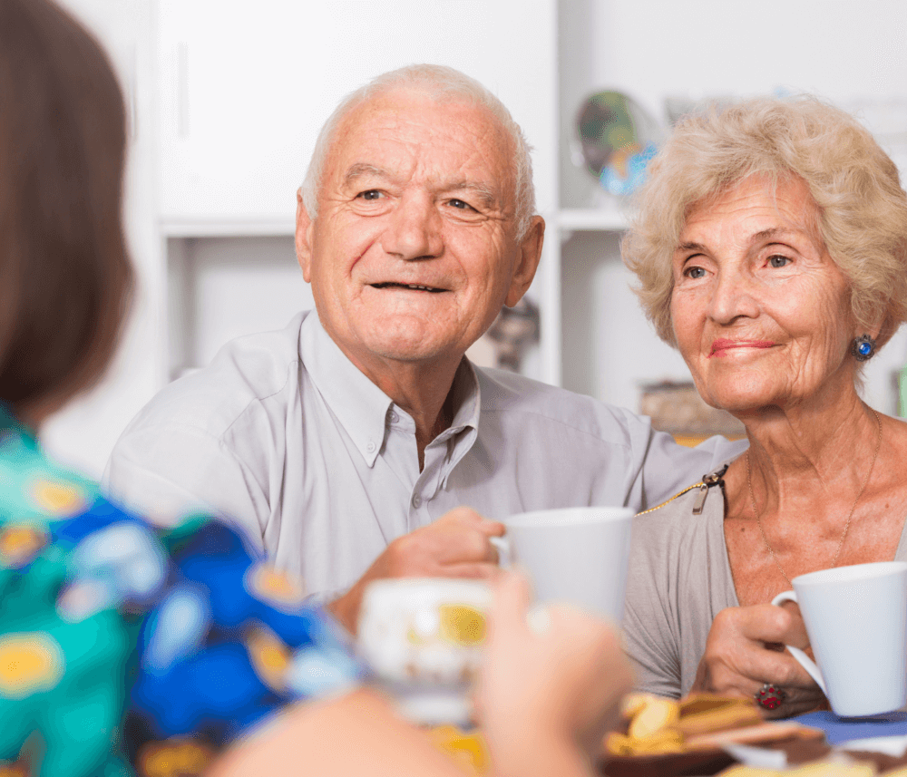 An elderly couple smiling and holding cups of coffee, sitting at a table with another person in the foreground. - Home Instead