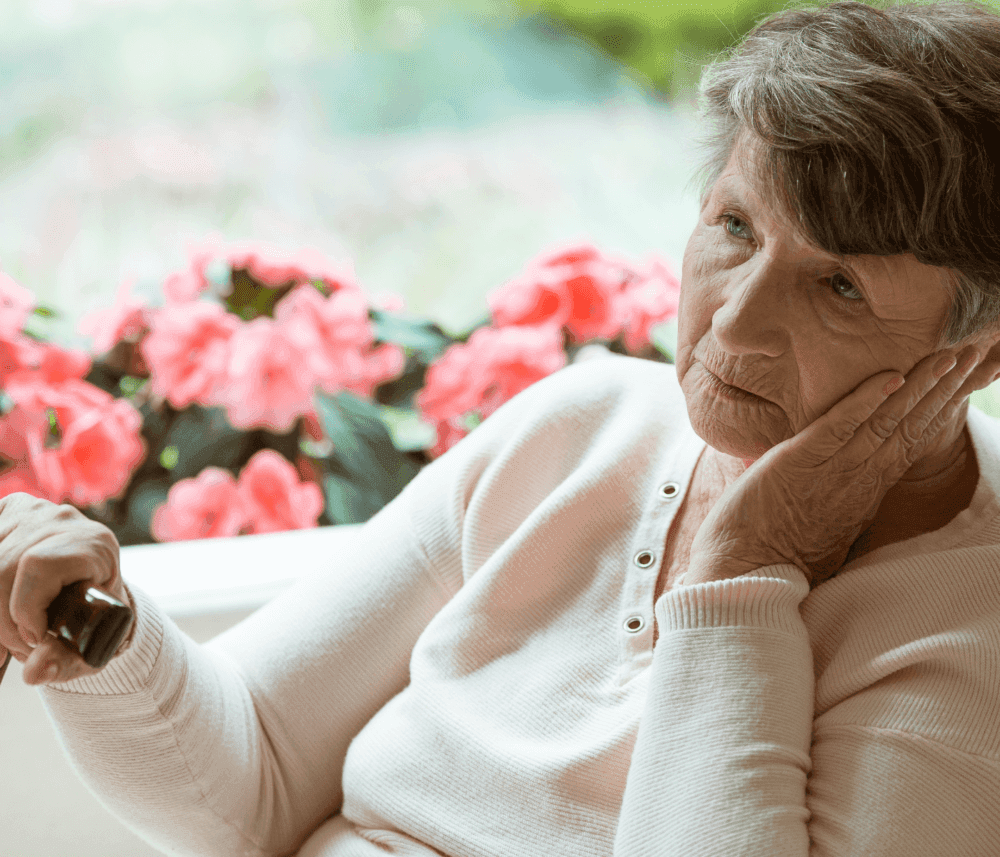 Older woman with gray hair, leaning on her hand, gazing contemplatively, with pink flowers in the background. - Home Instead