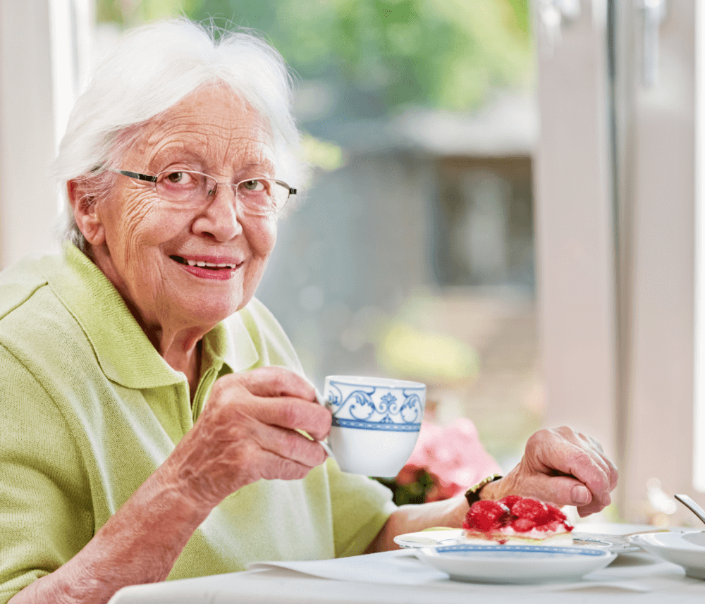 Elderly woman with glasses enjoying tea and dessert at a table, smiling at the camera. - Home Instead