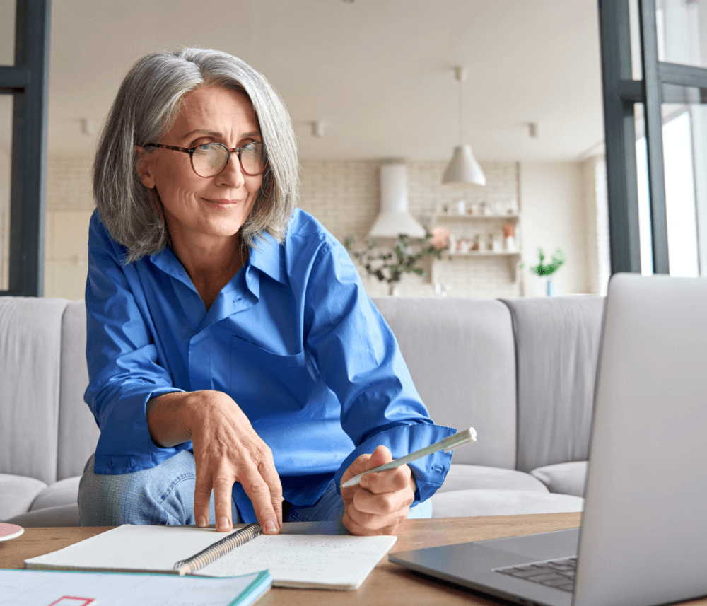 Older woman with glasses holding a pen, working on a laptop at home, with a notebook and a desk calendar. - Home Instead