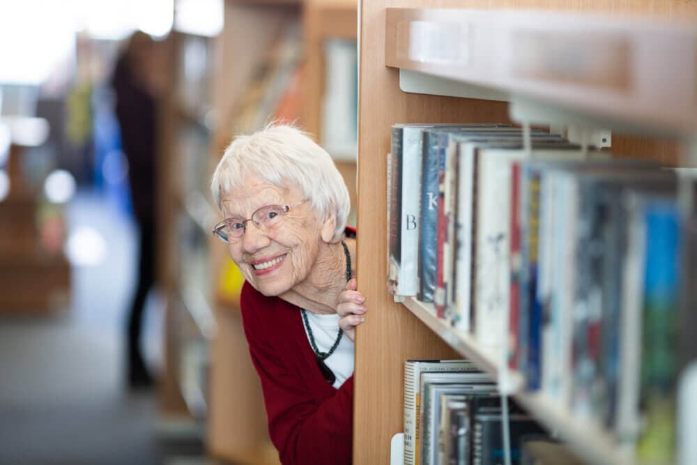 Elderly woman with glasses and white hair smiles while peeking around a bookshelf in a library. - Home Instead