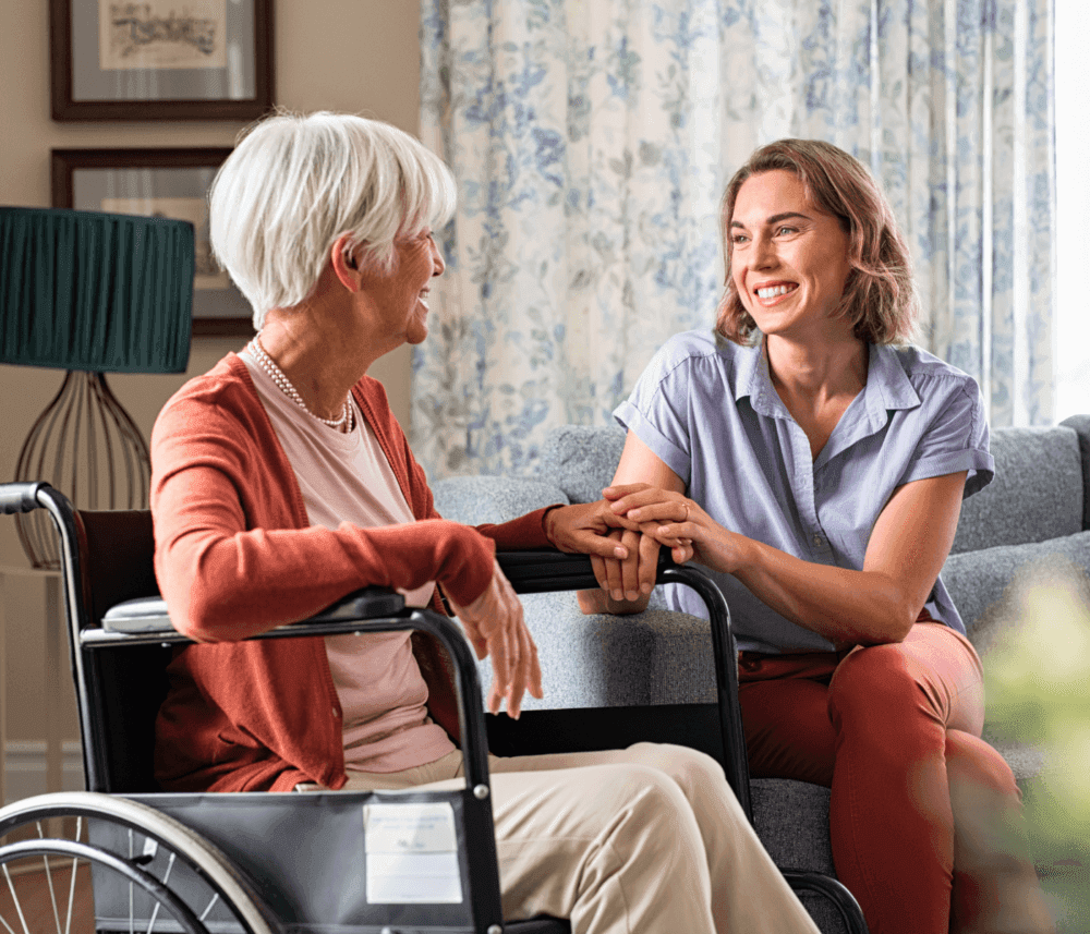 A smiling woman holds hands with an elderly woman in a wheelchair, sharing a warm moment in a cozy, sunlit room. - Home Instead