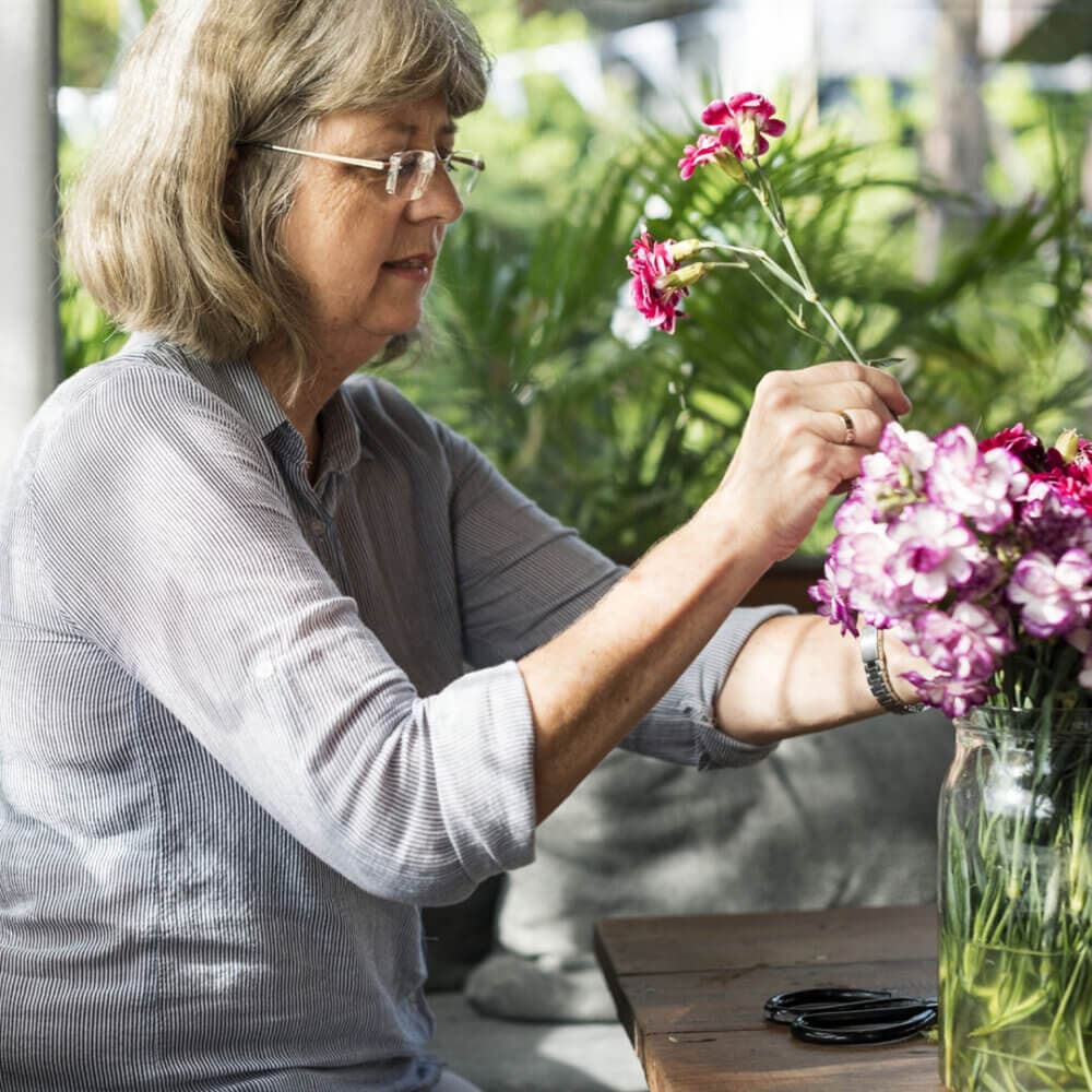 A woman arranges magenta flowers in a vase on a wooden table with greenery in the background. - Home Instead
