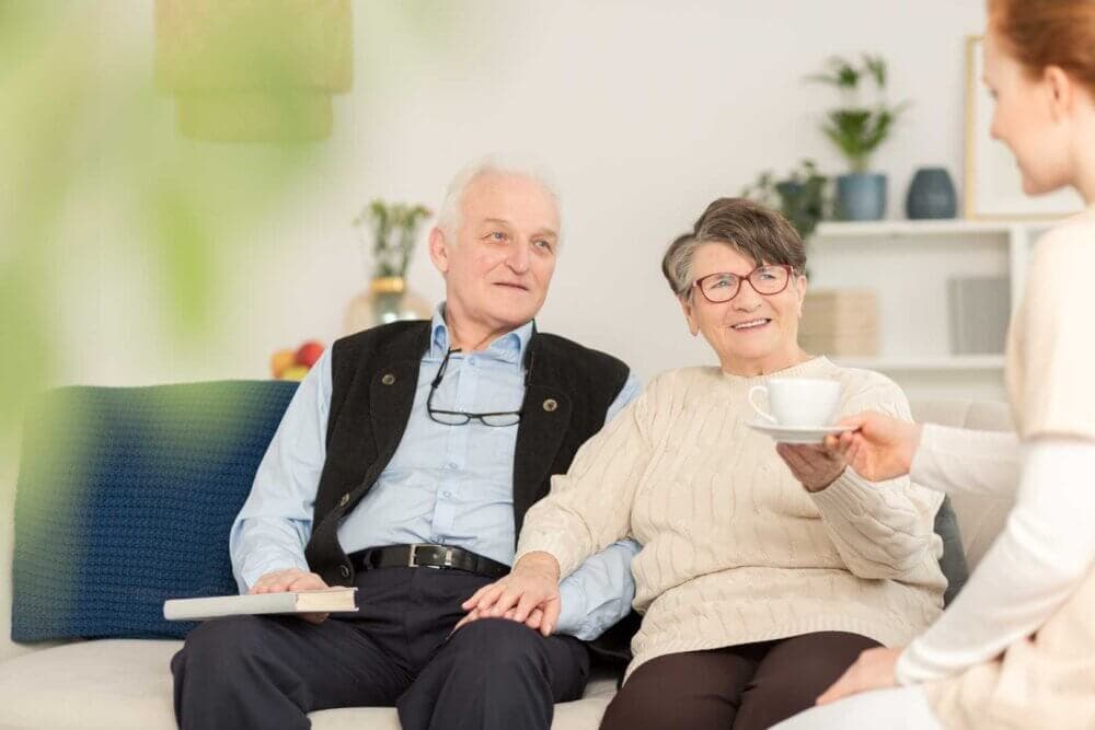 An elderly couple sits on a couch, smiling and talking to a young woman offering them tea. - Home Instead