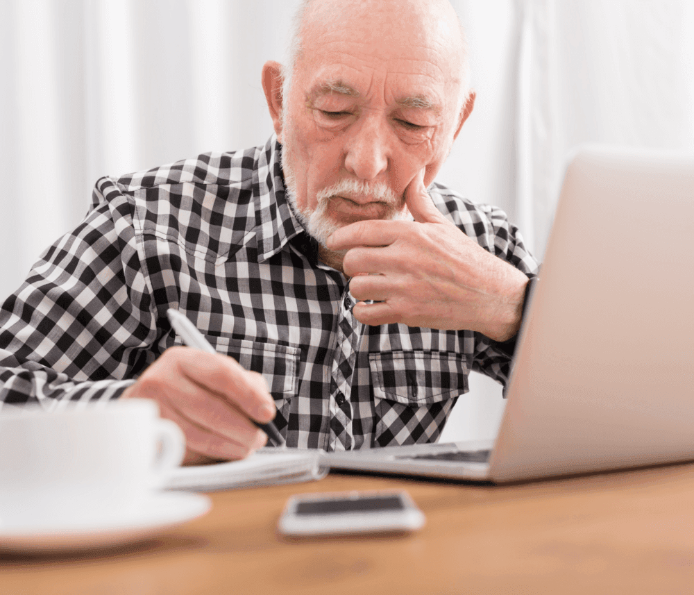 Elderly man in checkered shirt writing on paper, looking at laptop screen, with phone and coffee cup on table. - Home Instead