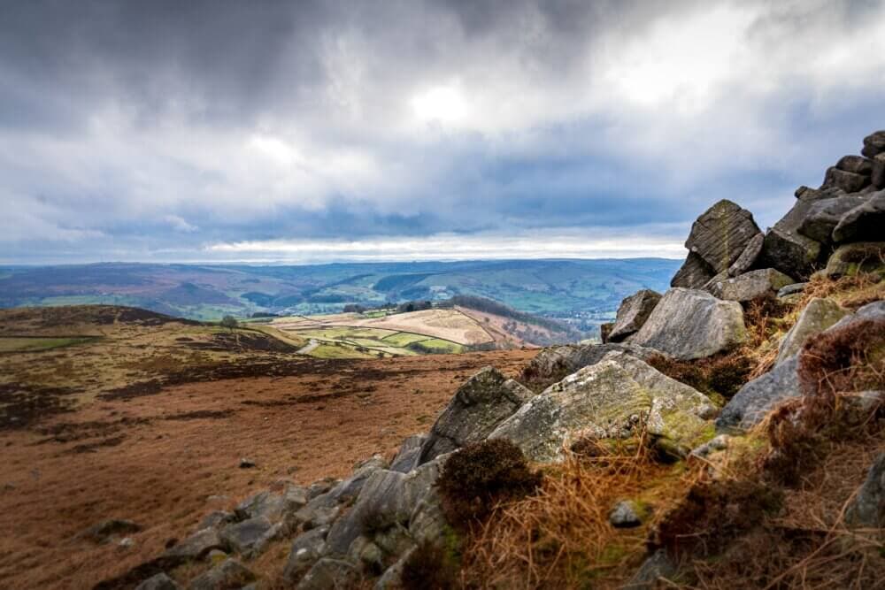 Rocky hillside with a vast view of a valley, hills, and fields under a cloudy sky. - Home Instead