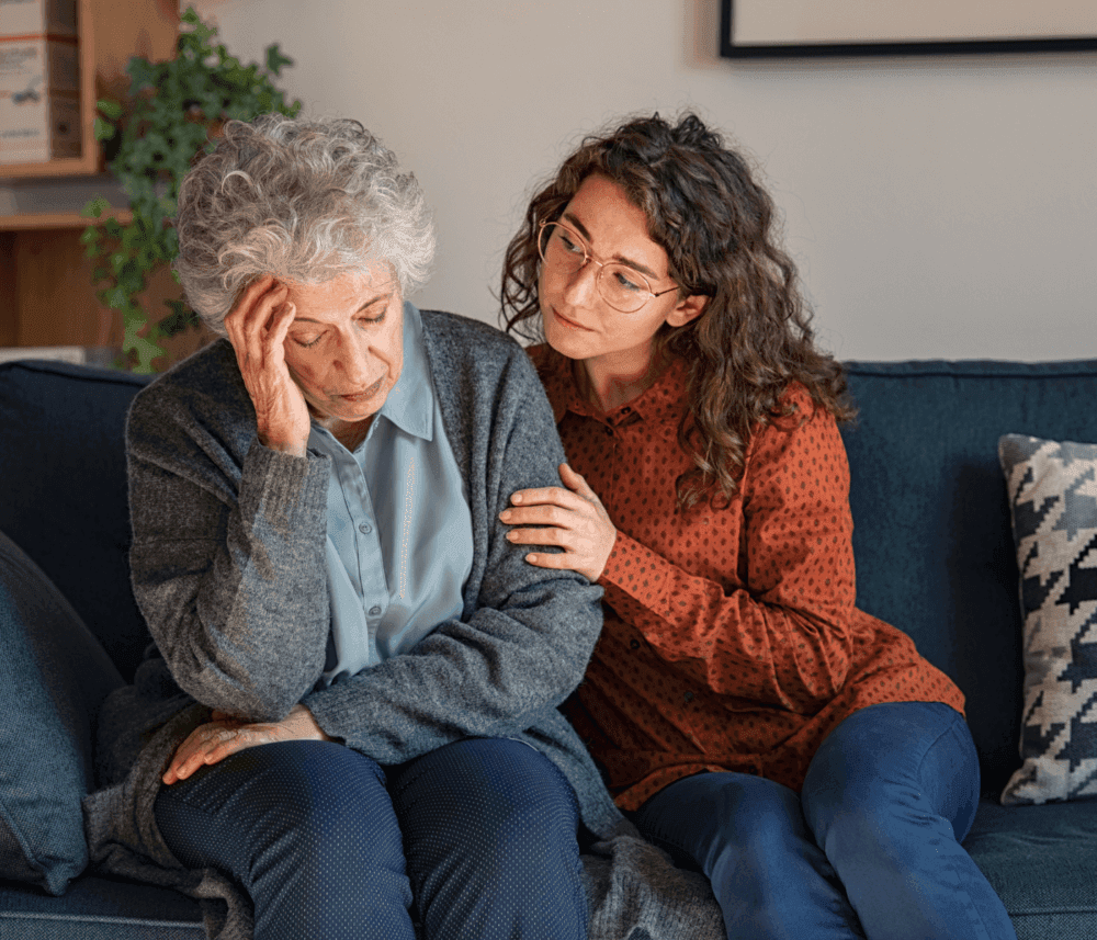 Young woman comforting an elderly woman sitting beside her on a couch, with the elderly woman looking distressed. - Home Instead