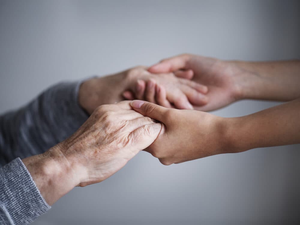 An elderly person and a younger person holding hands, symbolizing support and connection, against a grey background. - Home Instead