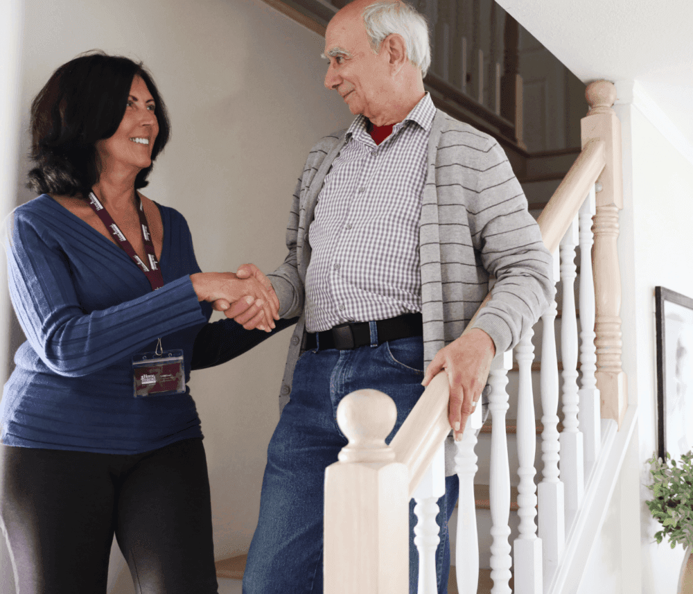 A woman with a name tag shakes hands with an elderly man standing on a staircase with a wooden handrail. - Home Instead