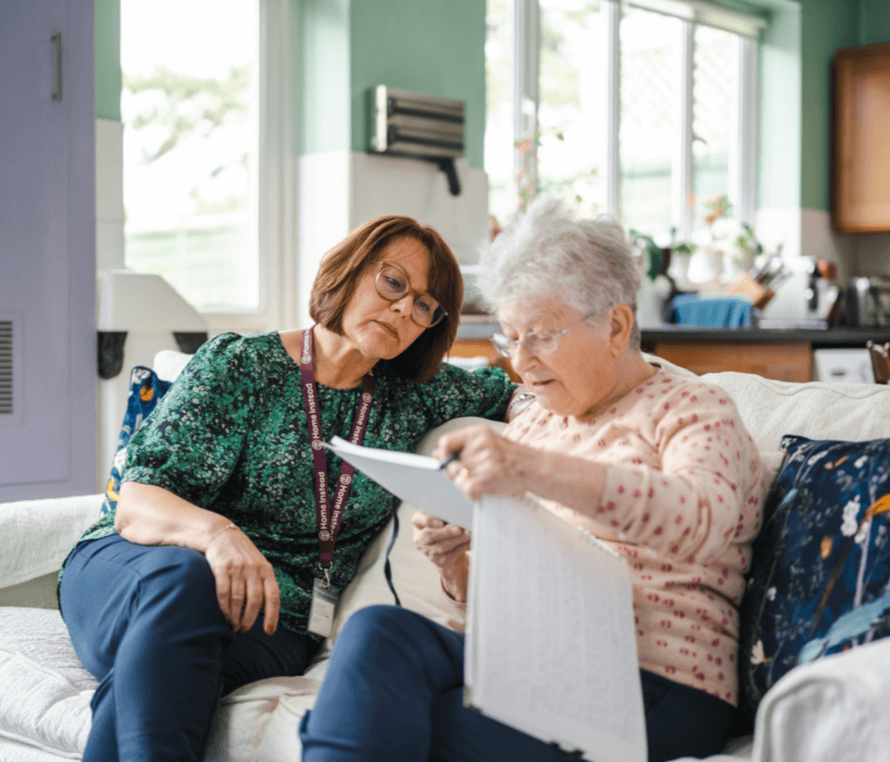 Two women sitting on a couch, one reading a document while the other looks on attentively. - Home Instead