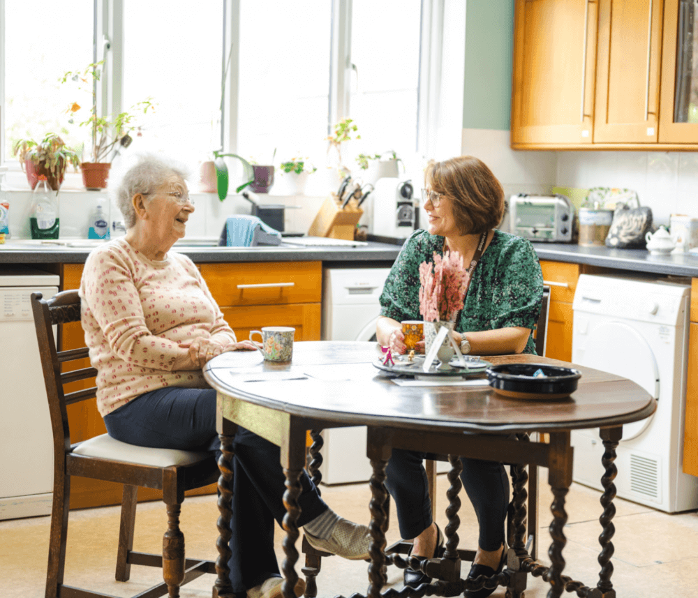Two women sitting at a kitchen table, smiling and having a conversation with coffee mugs in front of them. - Home Instead