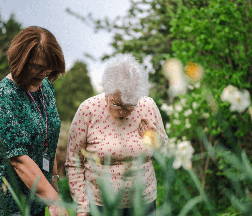 Two elderly women with grey hair, one in a green dress and the other in a polka-dot shirt, looking at plants. - Home Instead