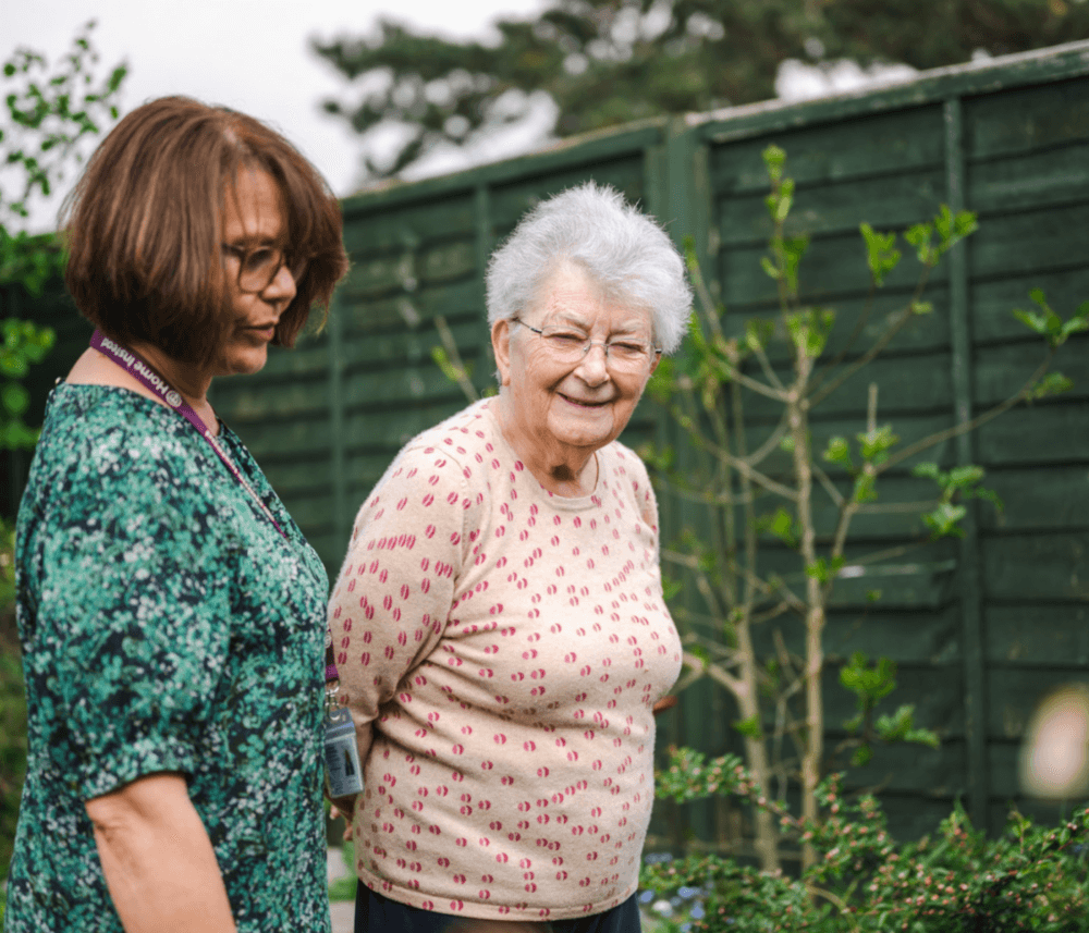 Two women, one older and one younger, walking together in a garden with green fencing in the background. - Home Instead