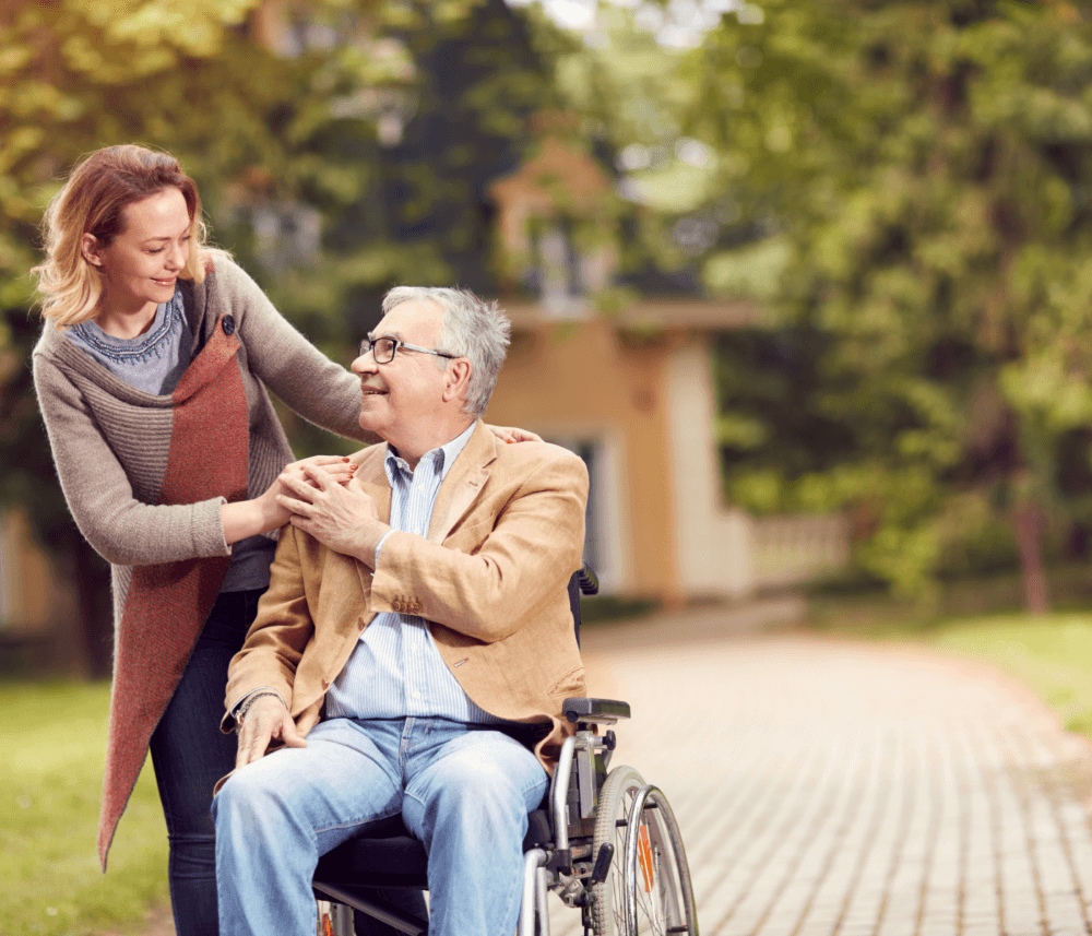 A woman smiles at a man in a wheelchair while gently touching his shoulder on a pathway in a park. - Home Instead