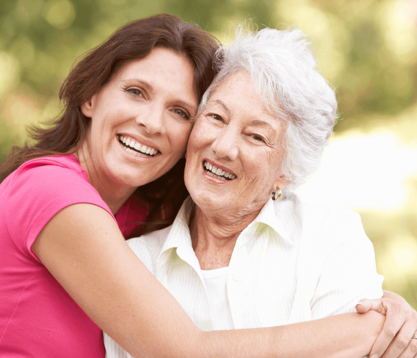 Two women, one younger and one older, smiling and embracing outdoors, with green trees in the background. - Home Instead