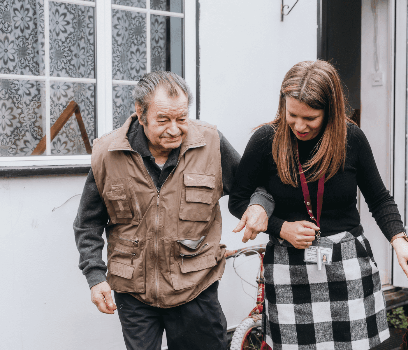 A woman assists a senior man by the arm as they walk outdoors near a white building with a window. - Home Instead