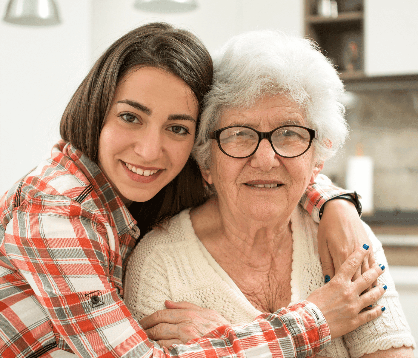 Young woman smiling and hugging an elderly woman who is also smiling, both in a bright indoor setting. - Home Instead