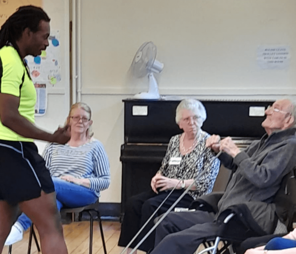 A man in athletic wear assists an elderly man with resistance bands as two women watch. - Home Instead