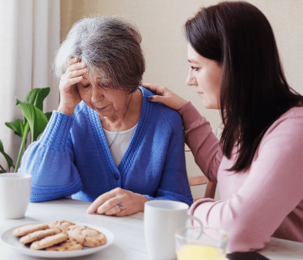 A young woman comforts an elderly woman sitting at a table with cookies and drinks, the elderly woman looks distressed. - Home Instead