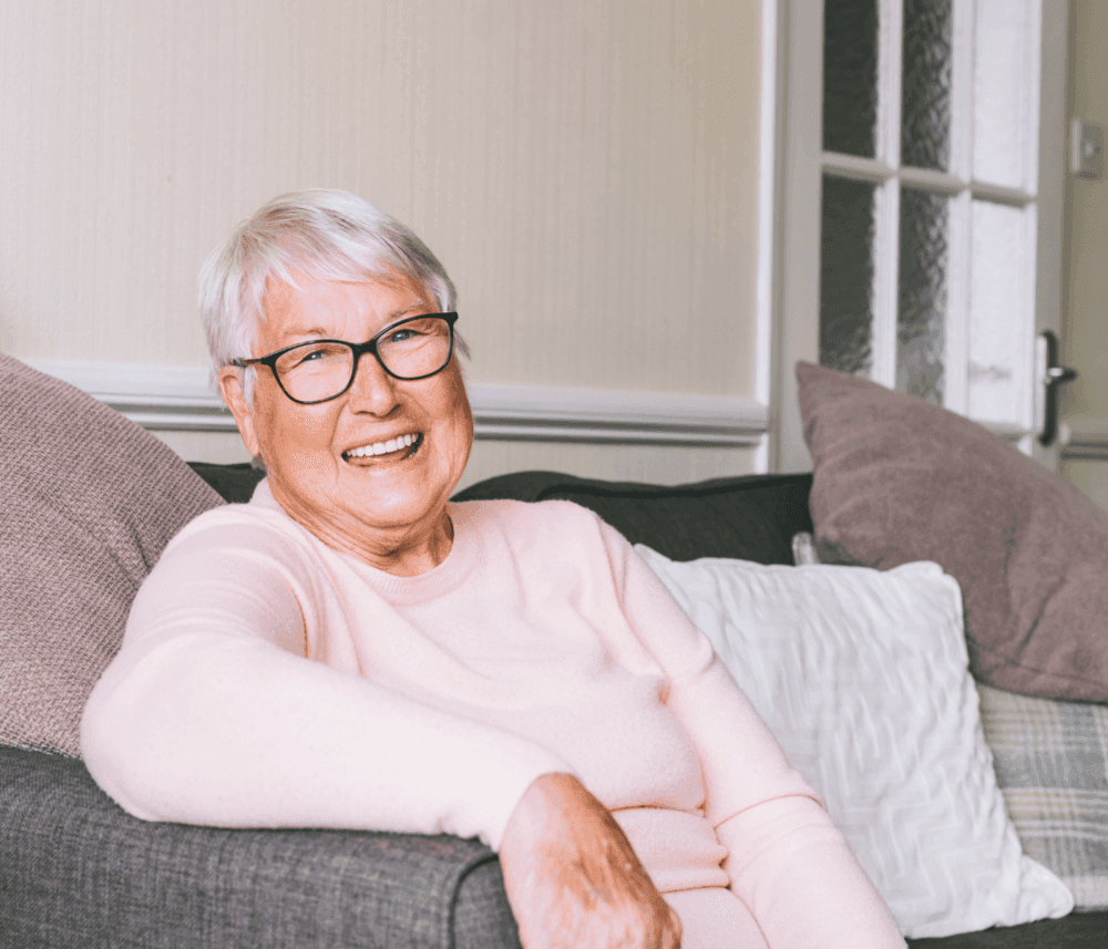Elderly woman with short white hair and glasses, smiling and sitting on a sofa, with pillows in the background. - Home Instead