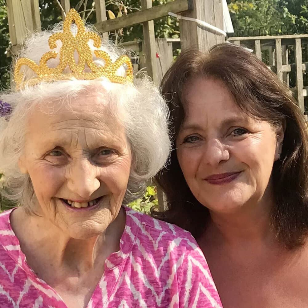Two women smiling; an older woman wears a golden tiara, and a younger woman with brown hair is beside her. - Home Instead