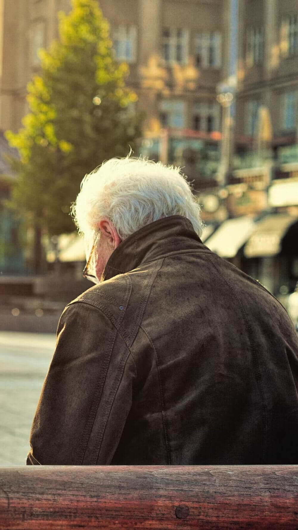 A person with grey hair, wearing a brown jacket, seated on an outdoor bench facing away from the camera. - Home Instead