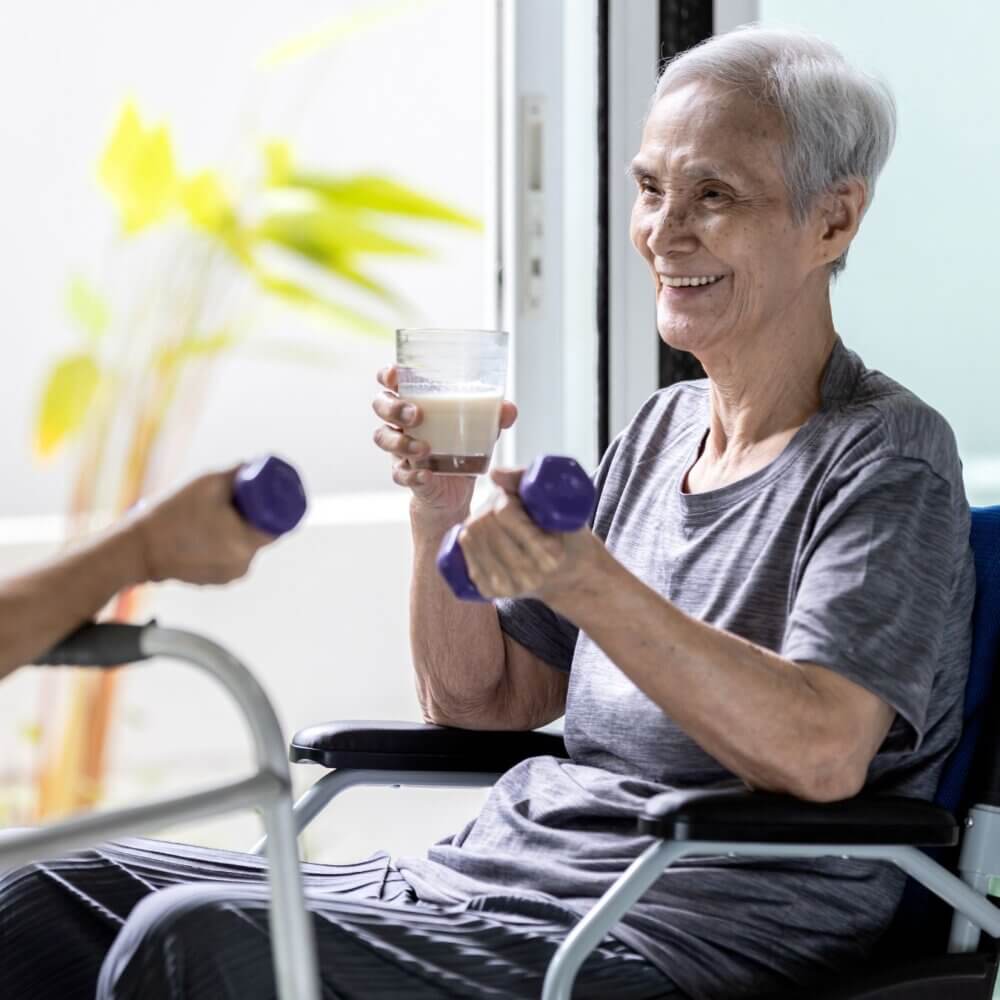 Elderly person in a wheelchair holding a dumbbell and a glass of milk, smiling, with a helping hand nearby. - Home Instead
