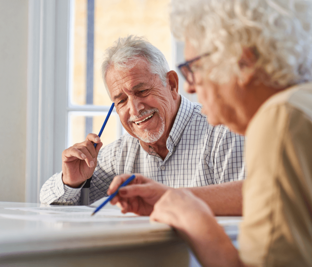 Two elderly people, one with glasses, are sitting at a table, smiling and holding pens, engaged in an activity. - Home Instead