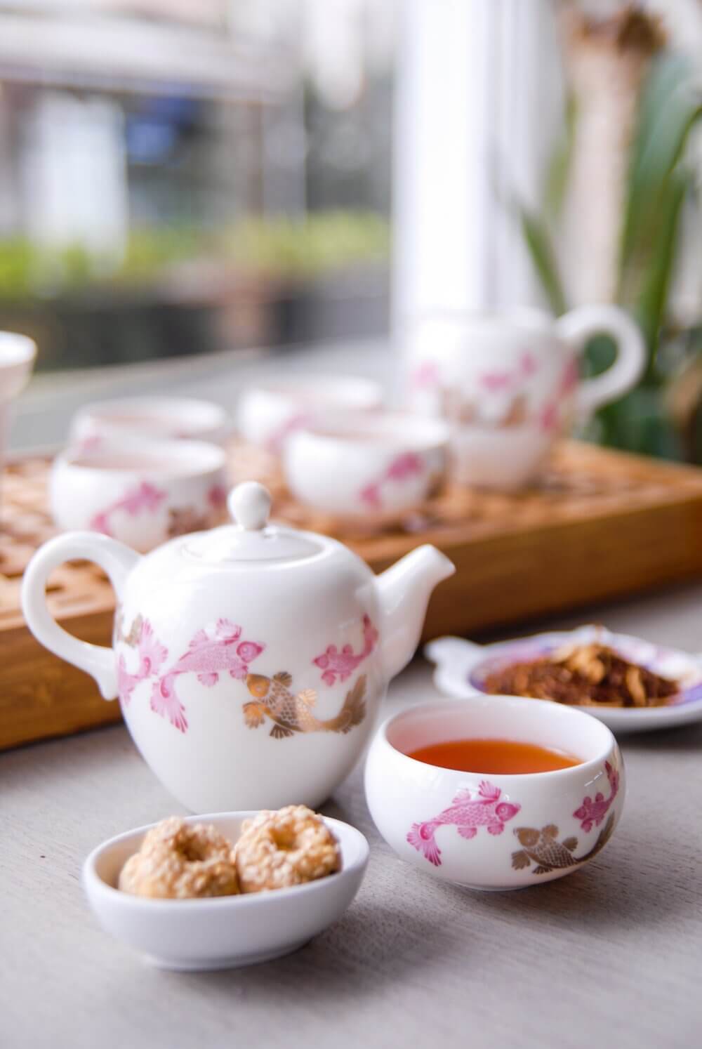 A floral teapot and teacup filled with tea next to biscuits, with more teacups in the background on a wooden tray. - Home Instead