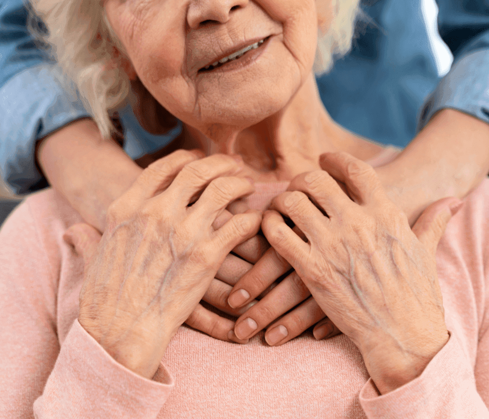 An elderly woman smiles gently as younger hands rest on her shoulders, symbolizing care and connection. - Home Instead
