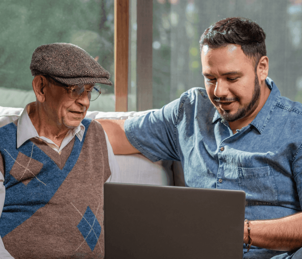 An elderly man and a younger man sit on a couch, smiling while looking at a laptop together. - Home Instead
