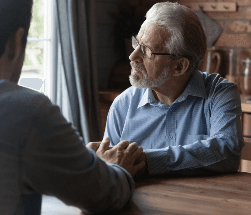Two people sitting at a table, one with gray hair and glasses, having a serious conversation while holding hands. - Home Instead