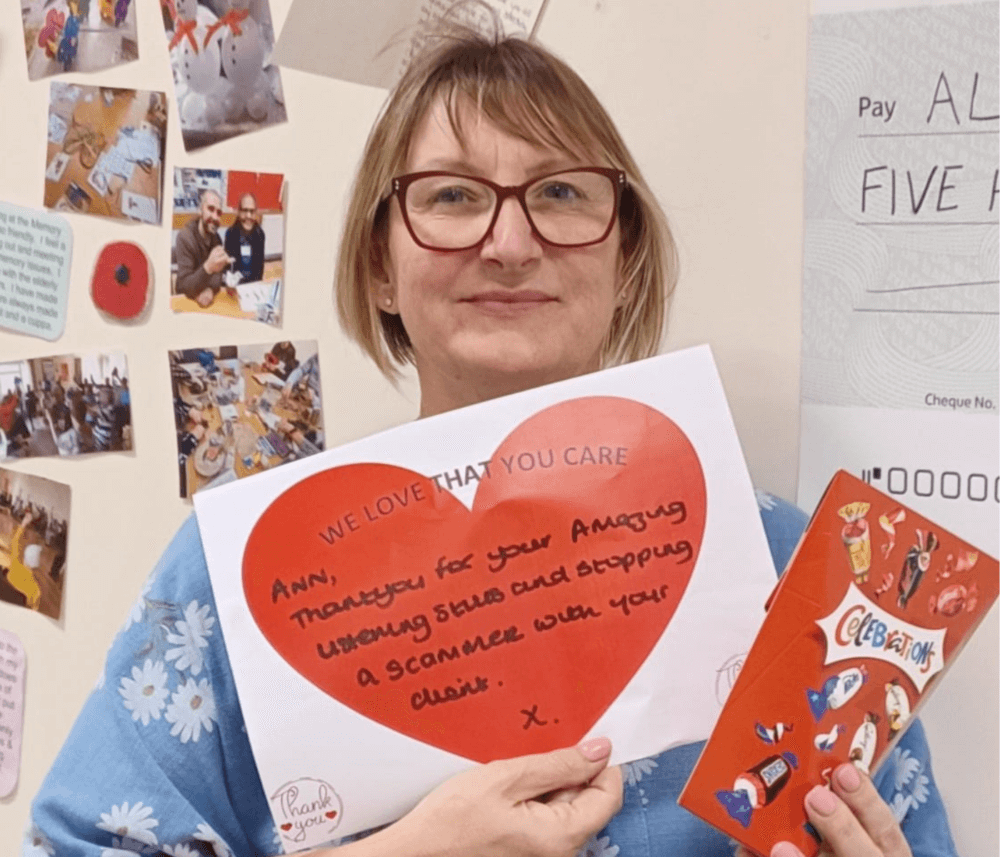 A woman with glasses holding a heart-shaped note and a "Celebrations" card, standing by a decorated wall. - Home Instead